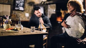 A rustic interior with stone walls, a wooden table with a glass bottle and a glass of water, and a person playing a stringed instrument. - Delina Mountain Resort (Anogia)