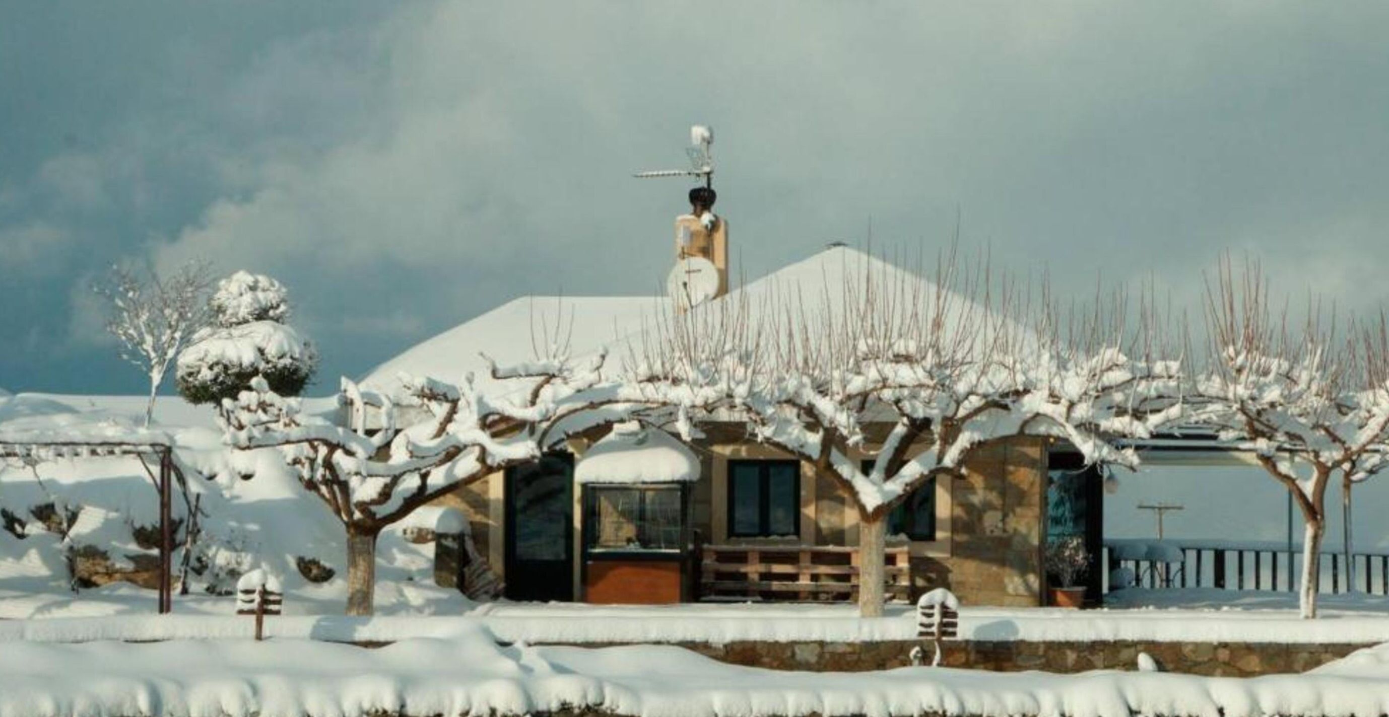 Una casa cubierta de nieve con una chimenea visible, un pequeño porche y un muro de piedra al frente.