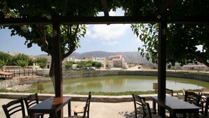 An outdoor seating area with tables and chairs, a pond, and buildings in the background. - Delina Mountain Resort (Anogia)