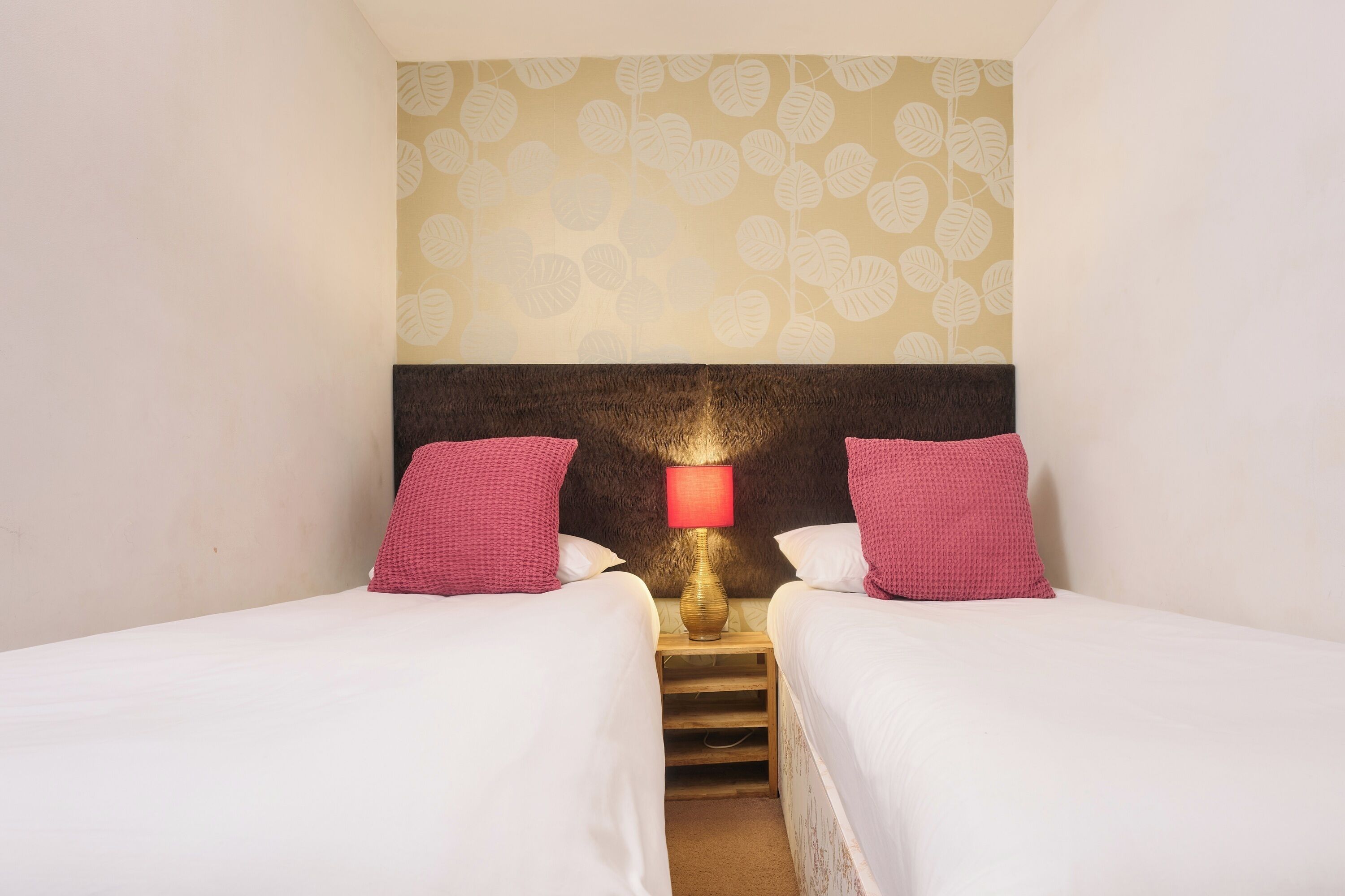 Habitación de hotel con dos camas, una mesita de noche con lámpara y un papel tapiz estampado.