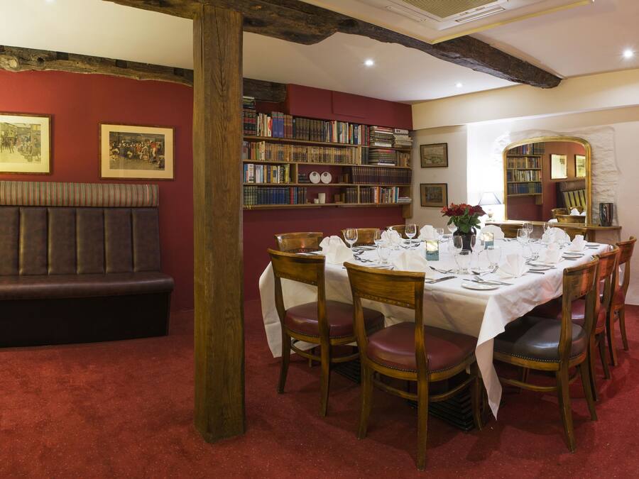 Un comedor con una mesa redonda dispuesta para cuatro personas, rodeada de sillas de madera. A la izquierda, una estantería llena de libros, y a la izquierda de la mesa, un banco con asiento acolchado.