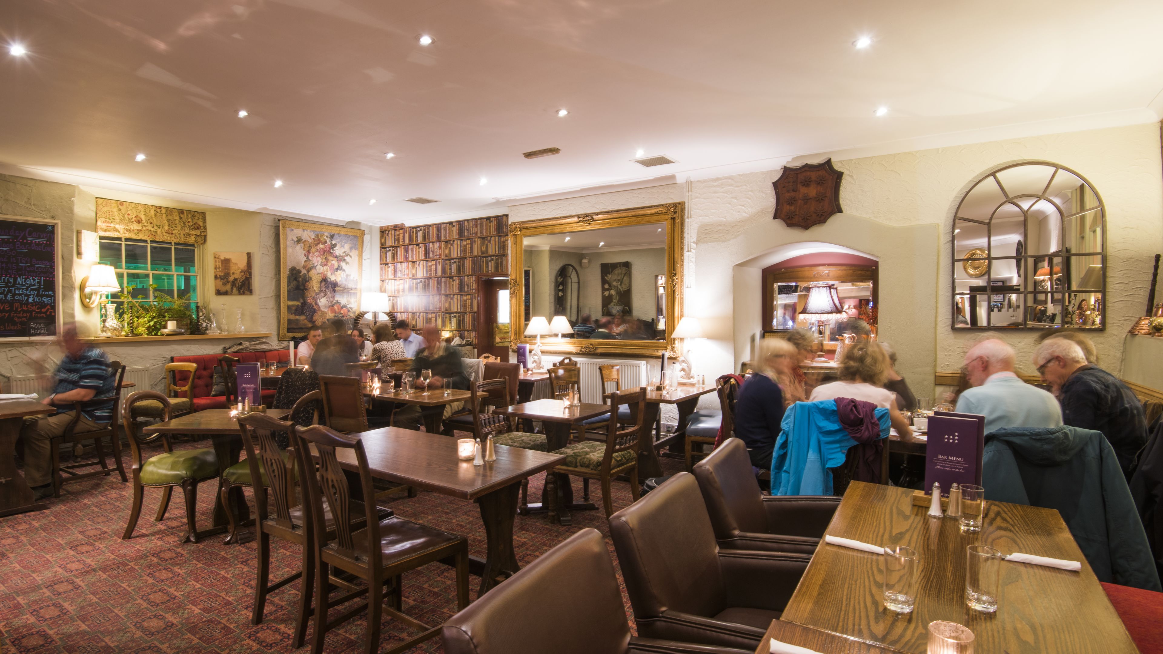 A cozy restaurant with wooden tables and chairs, a large mirror, and a bookshelf filled with books.