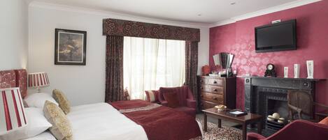 A hotel room with a large bed, a fireplace, a television mounted on the wall, and a desk with a lamp.
