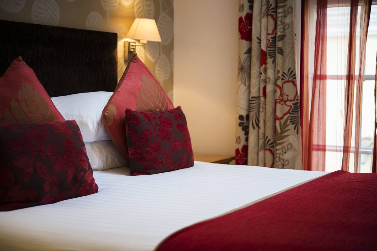 Un lit bien fait, agrémenté d’oreillers à motifs rouges et bruns, d’une couvre-lit blanche et d’un plaid rouge.