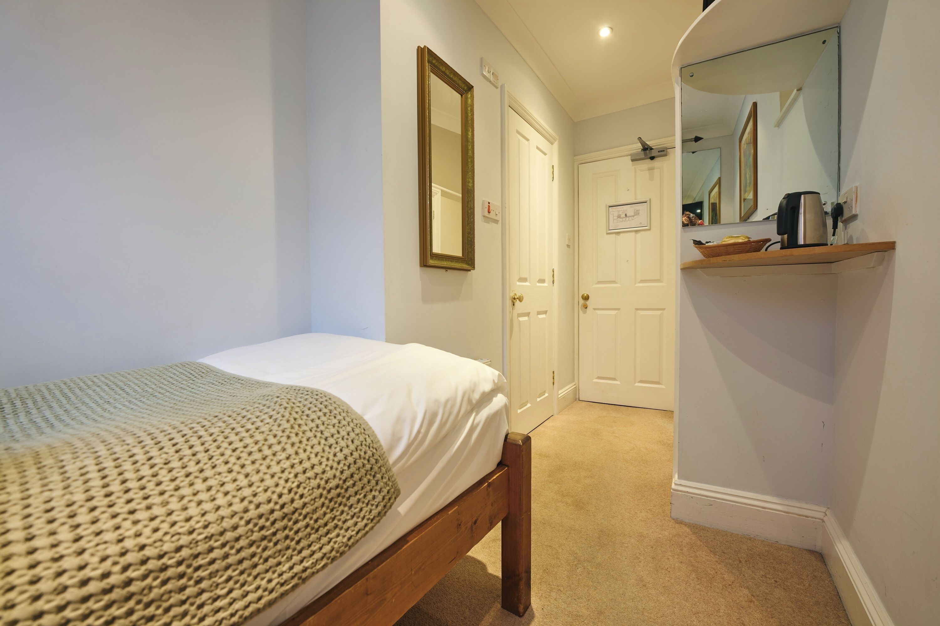 A bedroom with a single bed, a wooden headboard, a mirror, a shelf with a kettle, and a door leading to another room.
