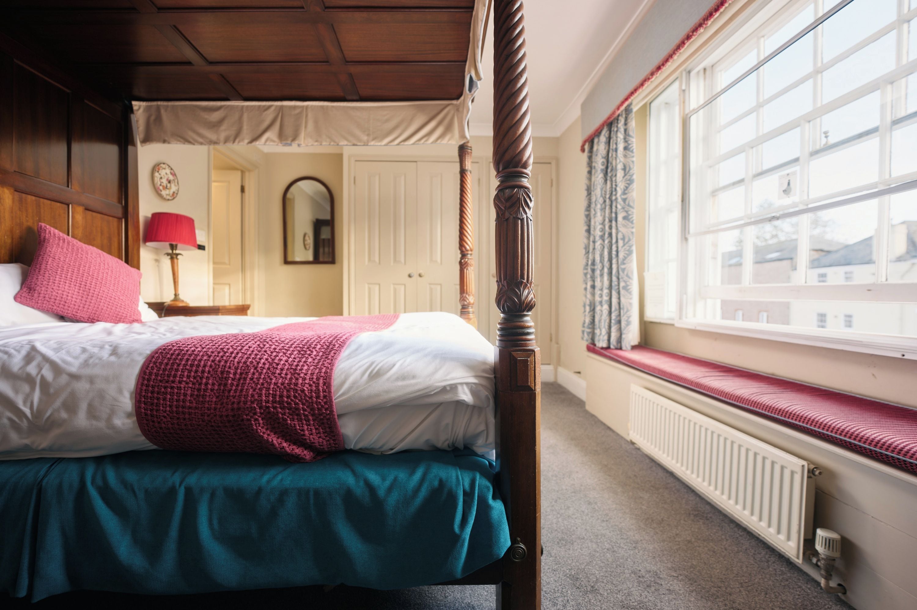 Una camera da letto con un letto a baldacchino, una seduta finestrino, un termosifone e uno specchio a parete.