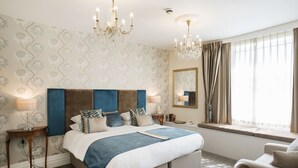 A bedroom with a large bed, two bedside tables, a chandelier, a window with curtains, and a mirror on the wall. - Royal Seven Stars Hotel (Totnes)