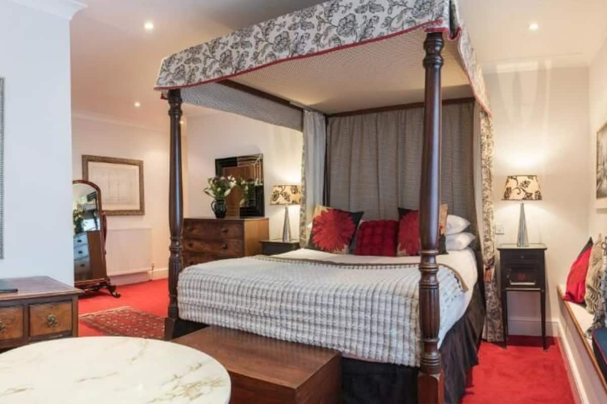 Un lit à baldaquin avec une couvre-lit à carreaux, une table de chevet avec une lampe, une chaise rouge et une table en marbre.