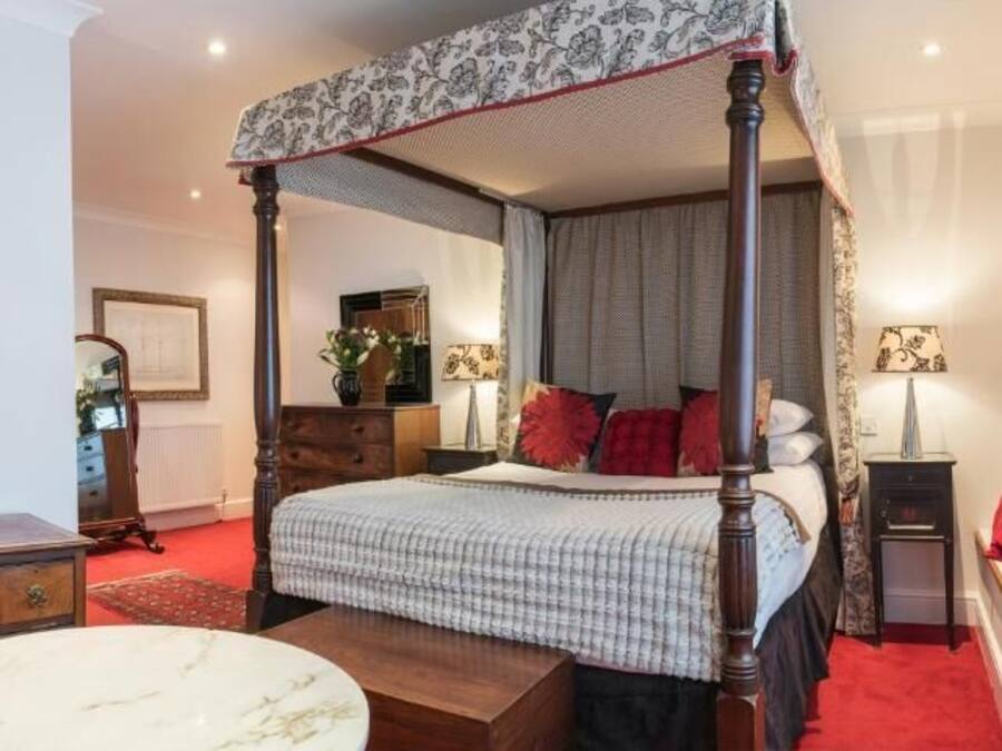 Una cama con dosel y edredón a cuadros, una mesita de noche con lámpara, una silla roja y una mesa de mármol.