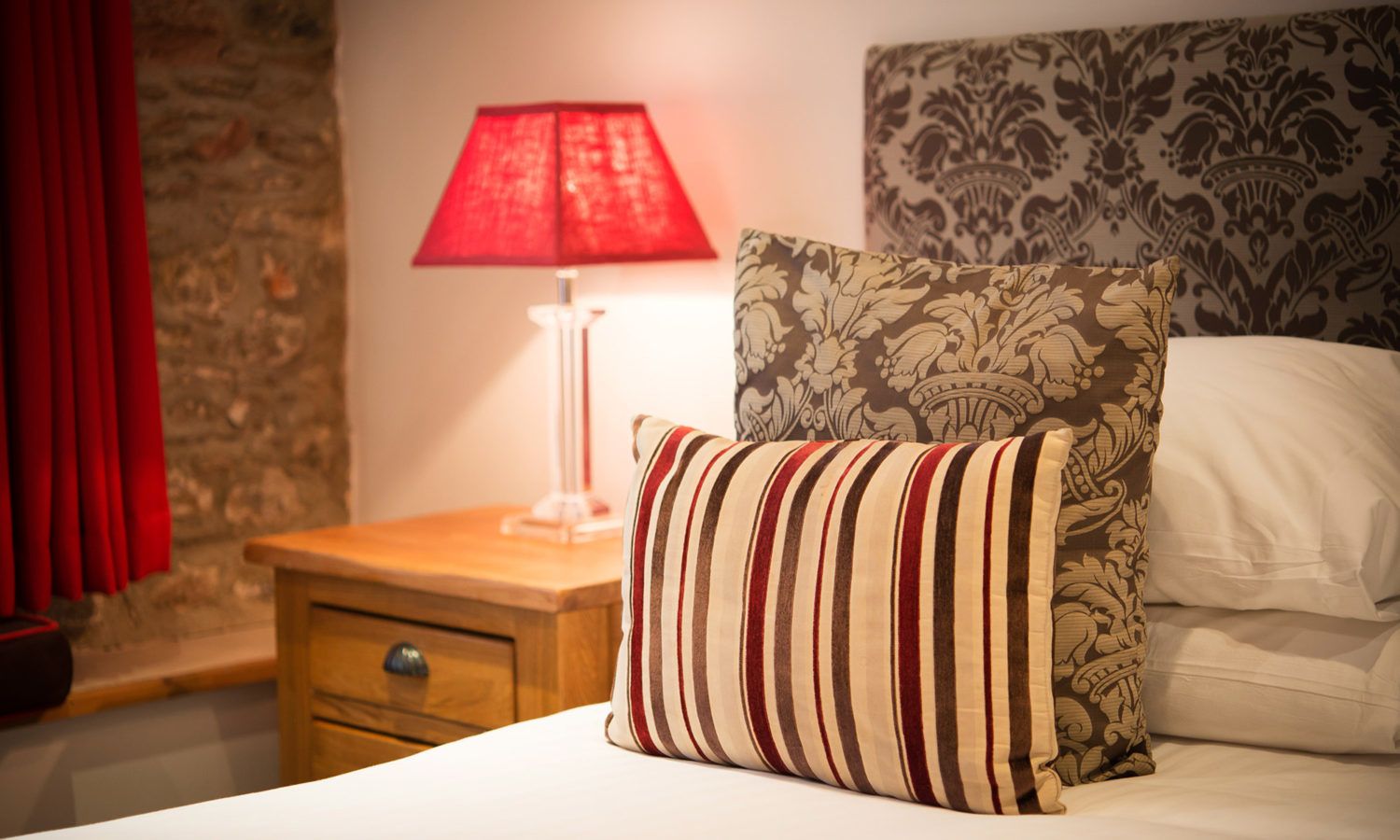 A neatly made bed with striped pillows, a red lamp on a bedside table, and a patterned headboard.