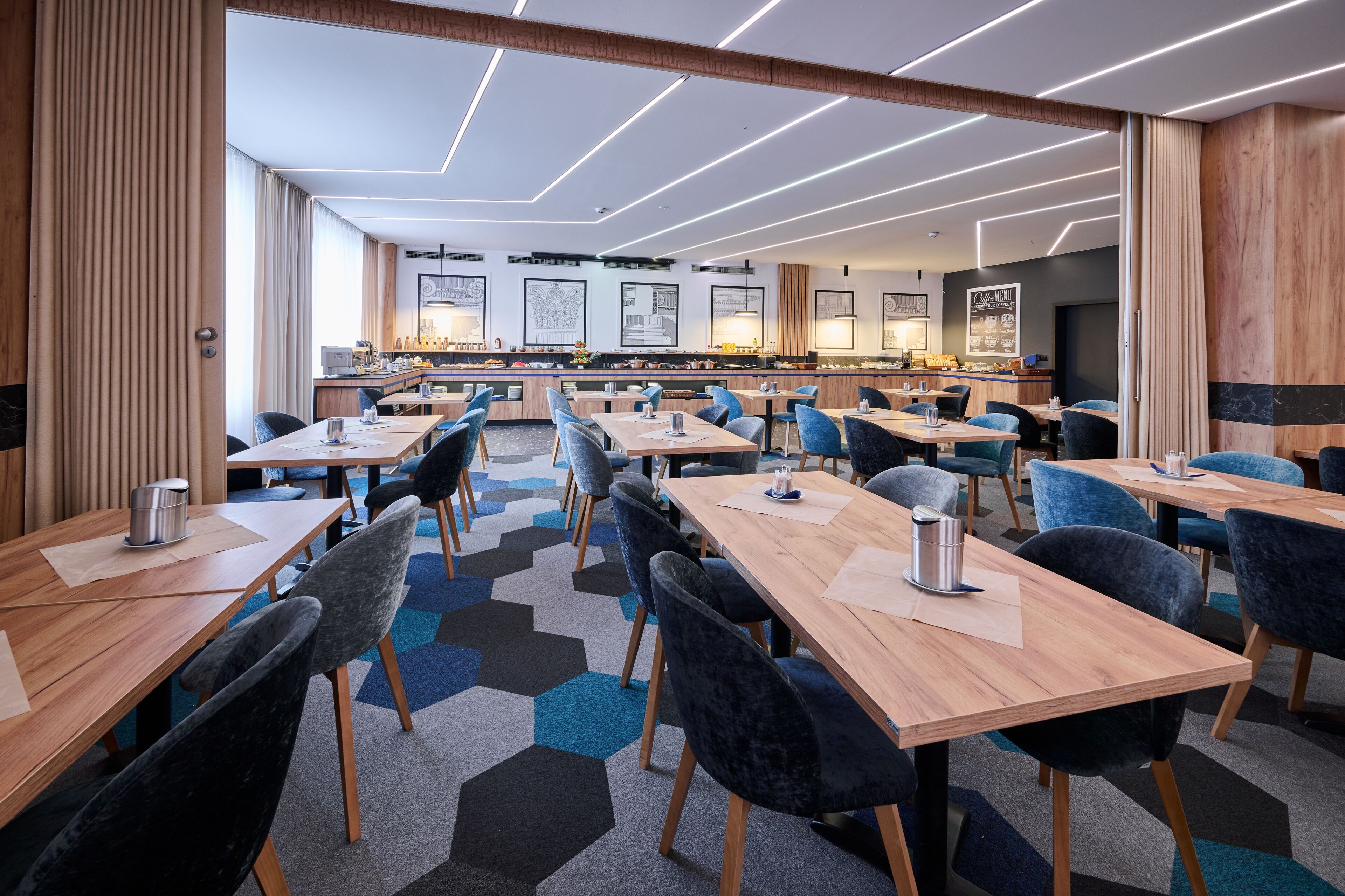 A modern hotel dining area with multiple tables and chairs, a kitchen in the background, and a geometric patterned carpet.