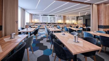 A modern hotel dining area with multiple tables and chairs, a kitchen in the background, and a geometric patterned carpet.