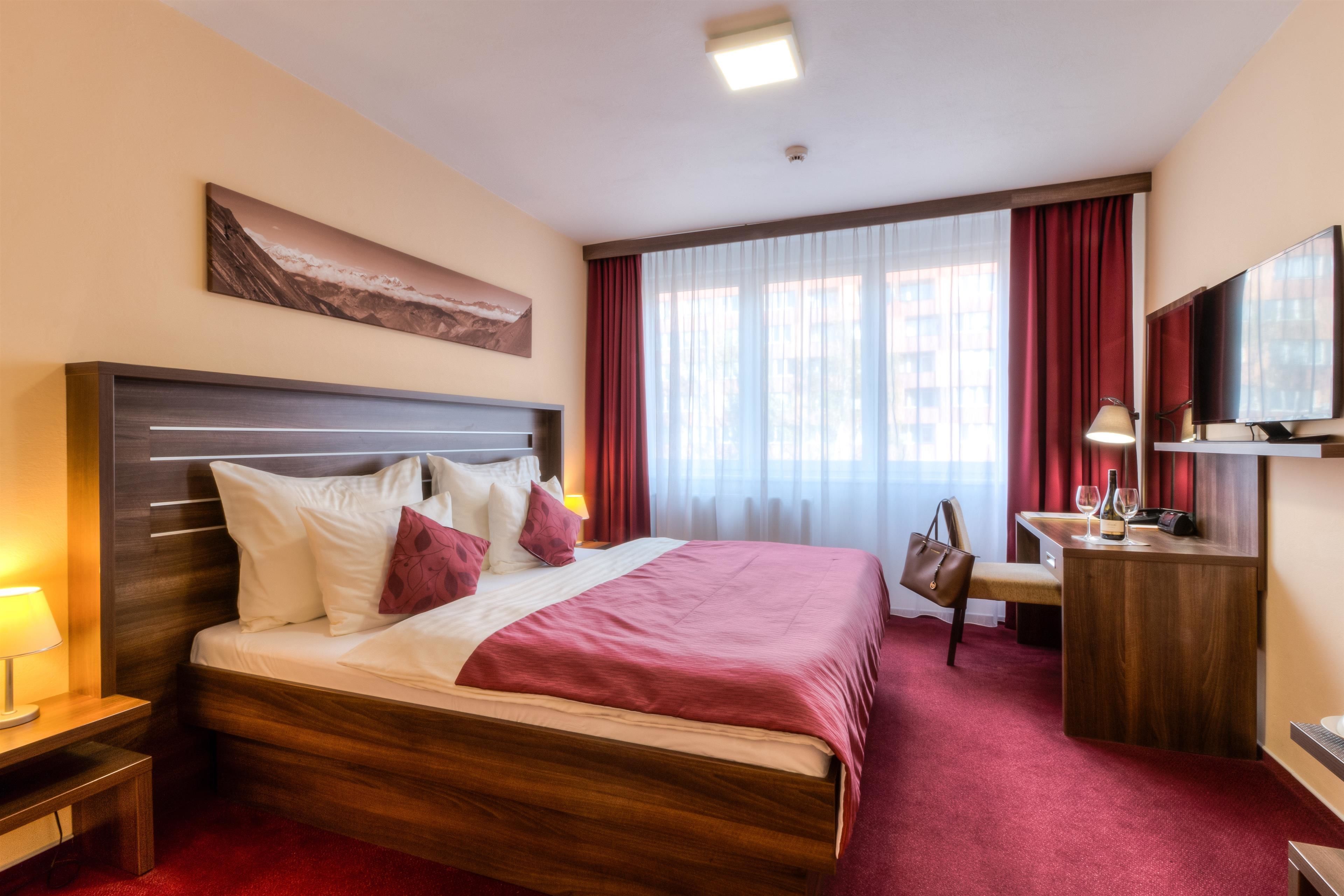 A hotel room with a large bed, a desk with a chair, a TV, and a window with curtains.