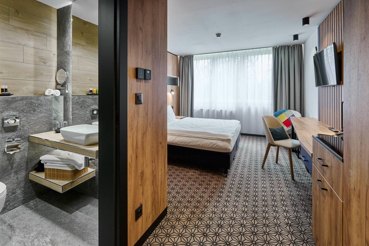 A hotel room with a wooden wall, a bed, a TV, a desk, and a bathroom with a sink and mirror.
