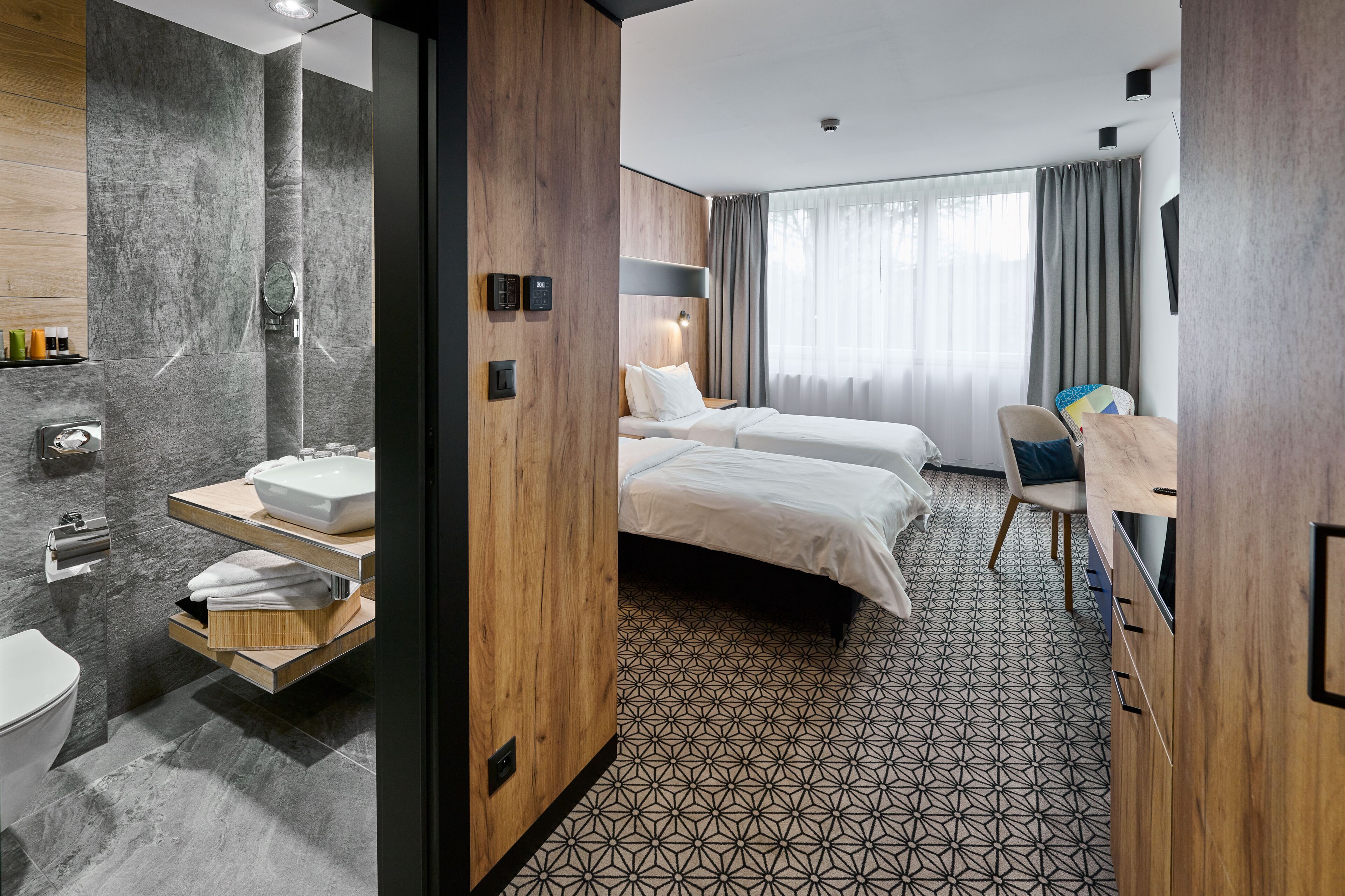 Habitación de hotel moderna con una cama grande, un escritorio, una silla, un baño con lavamanos e inodoro, y un ventanal con cortinas.