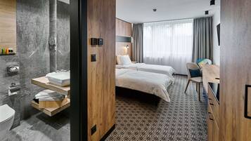 Habitación de hotel moderna con una cama grande, un escritorio, una silla, un baño con lavamanos e inodoro, y un ventanal con cortinas.