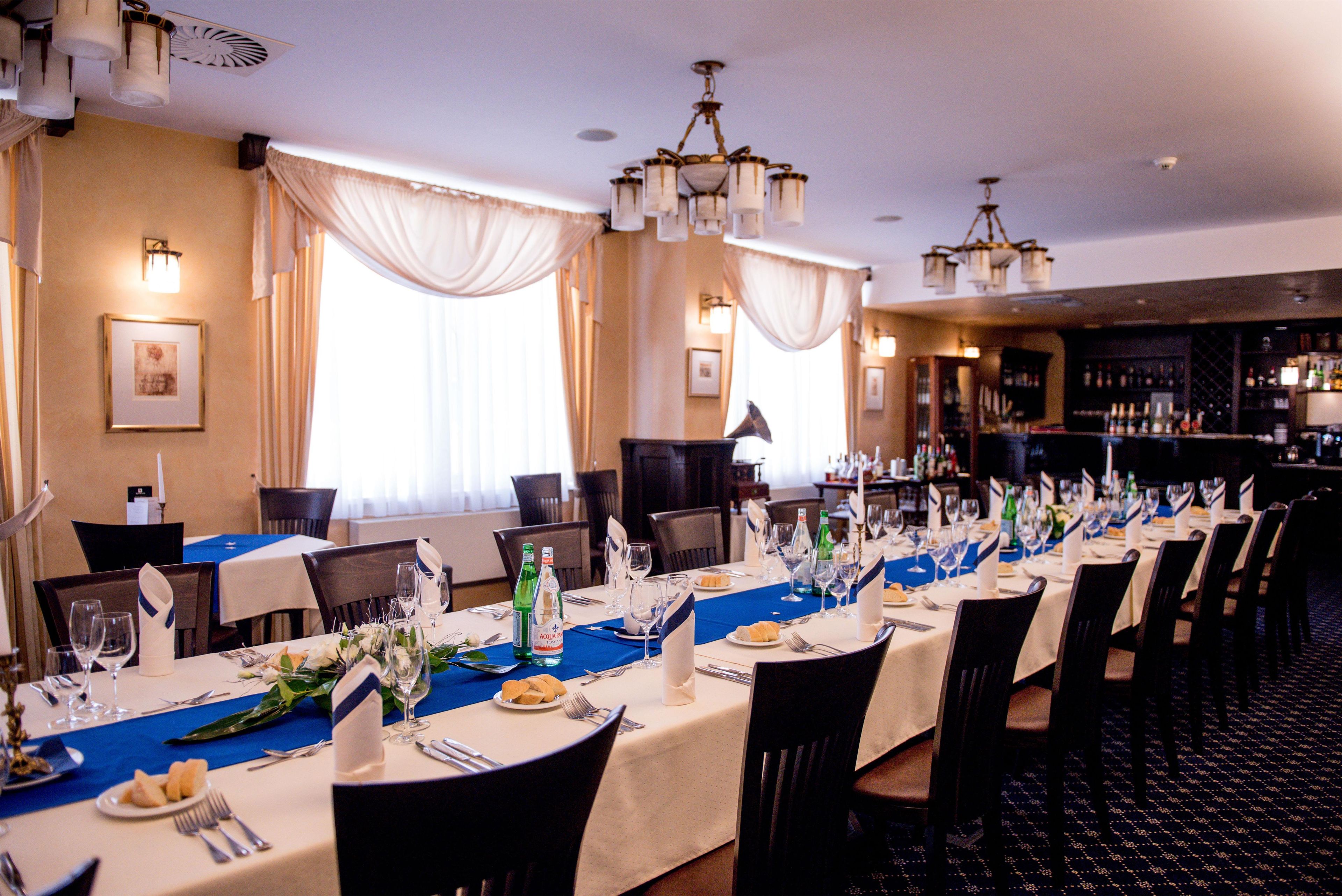 A formal dining room with long tables set for multiple guests, featuring a blue table runner, white tablecloths, and a bar area in the background.