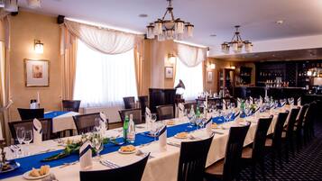 A formal dining room with long tables set for multiple guests, featuring a blue table runner, white tablecloths, and a bar area in the background.