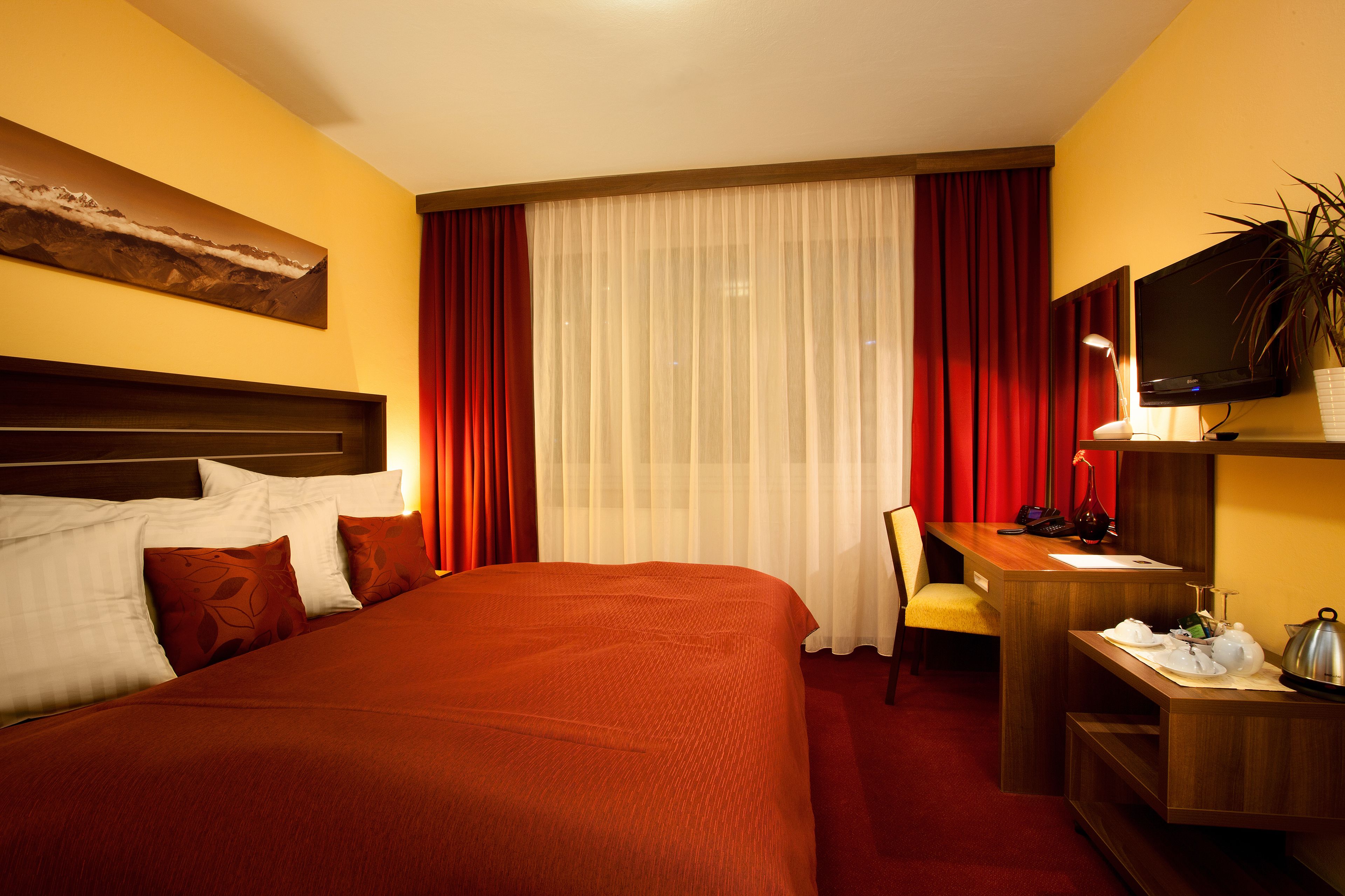 Quarto de hotel com uma cama grande, uma escrivaninha com cadeira, televisão e uma planta.