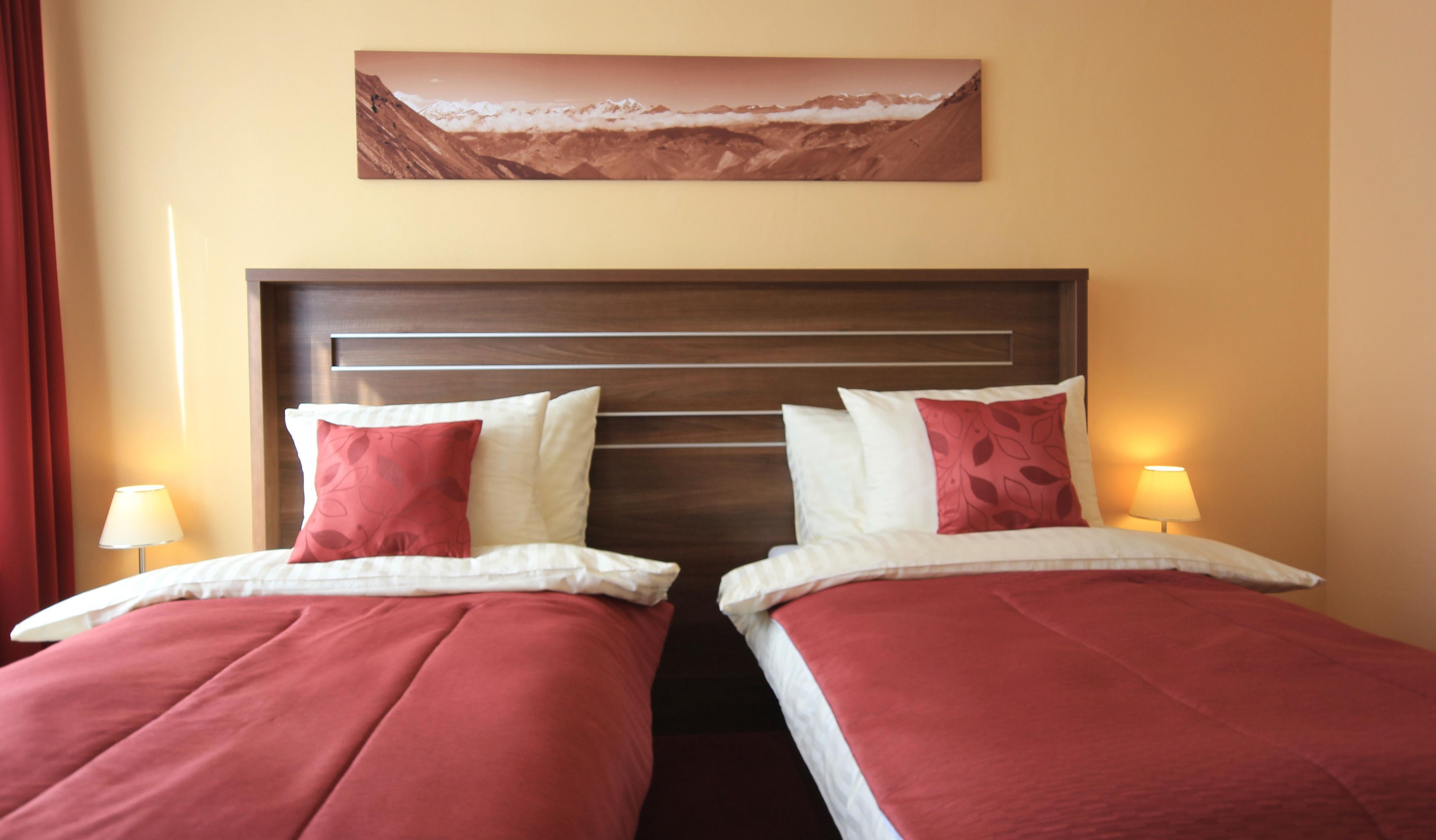 Habitación de hotel con dos camas, cada una con ropa de cama roja y blanca, cabeceros de madera y un cuadro de un paisaje montañoso sobre las camas.