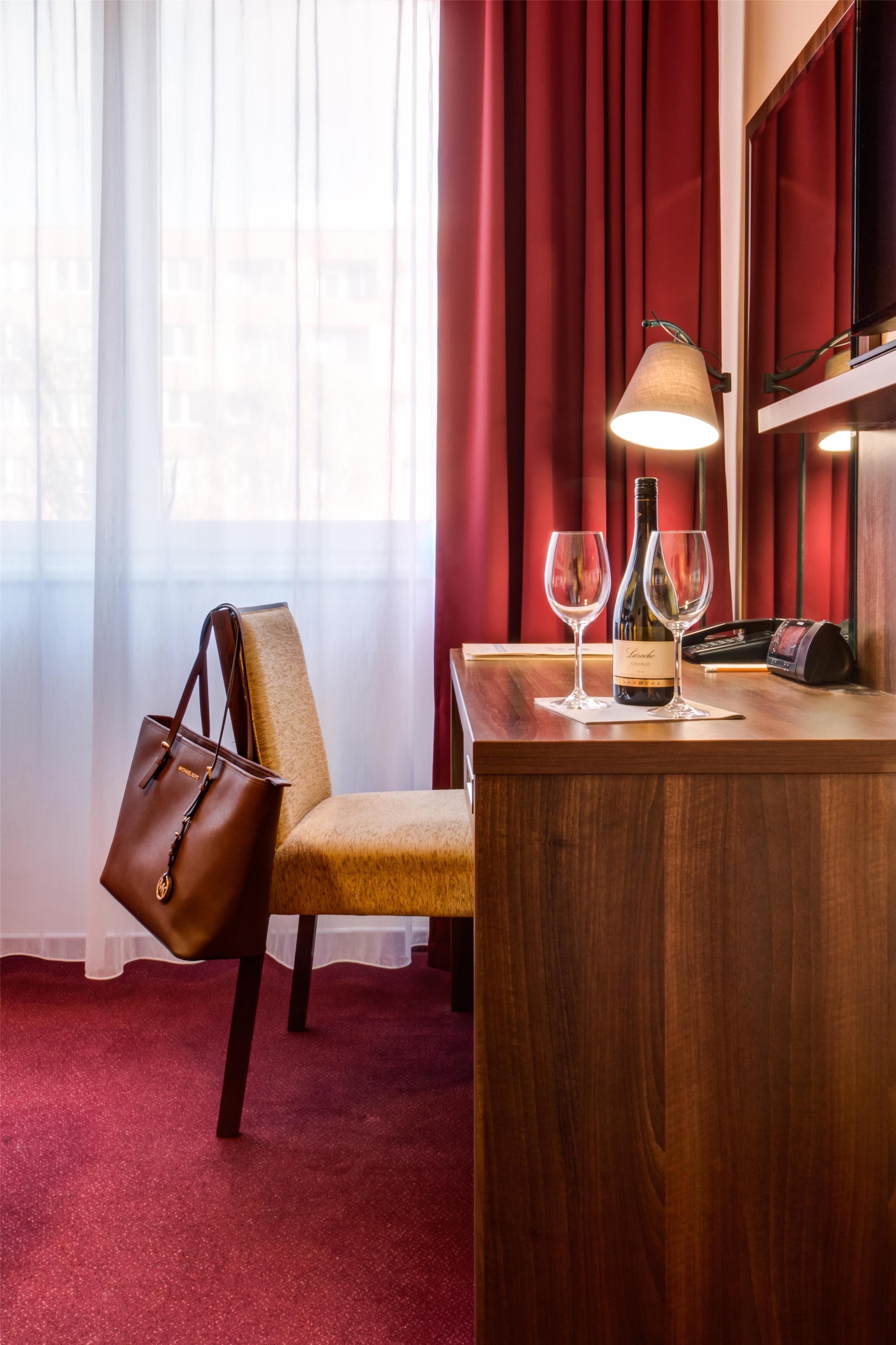 A hotel room with a wooden desk, a chair, a handbag, a bottle of wine, and a wine glass.