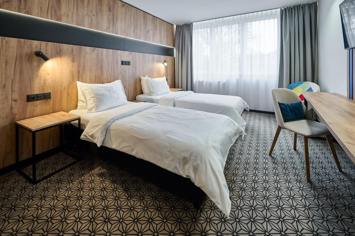 A hotel room with two beds, a wooden headboard, a desk with a chair, and a large window with curtains.