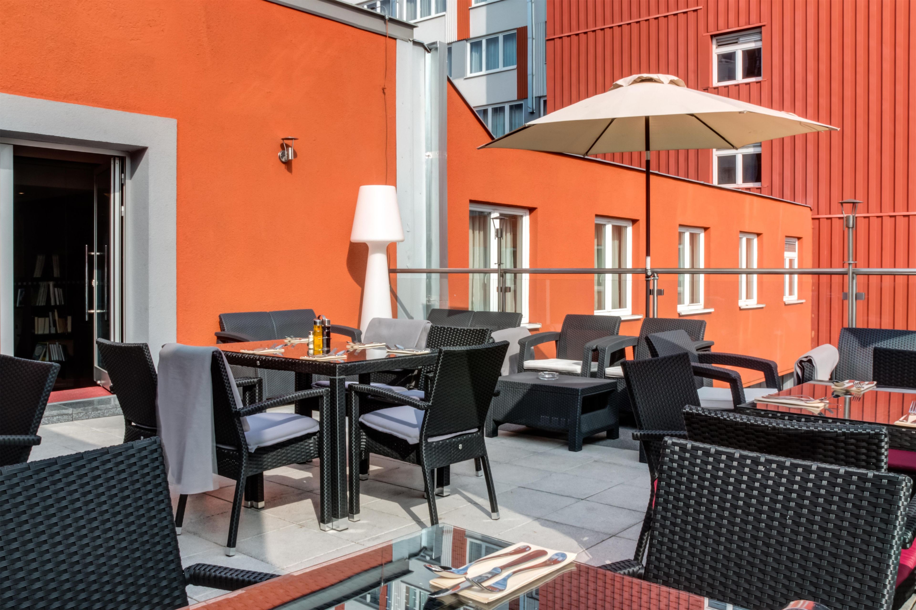 An outdoor seating area with wicker furniture and a glass table, overlooking a red building with multiple windows.