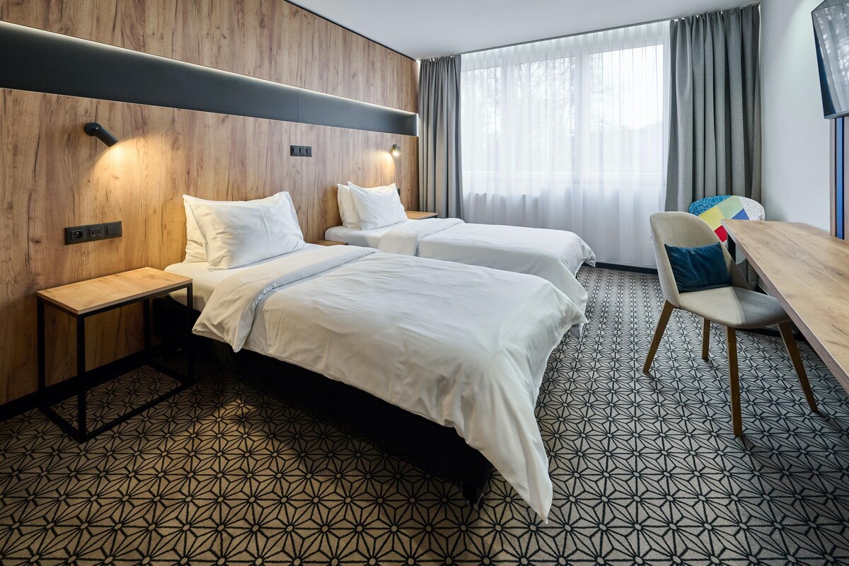 A hotel room with two beds, a wooden headboard, a desk with a chair, and a large window with curtains.