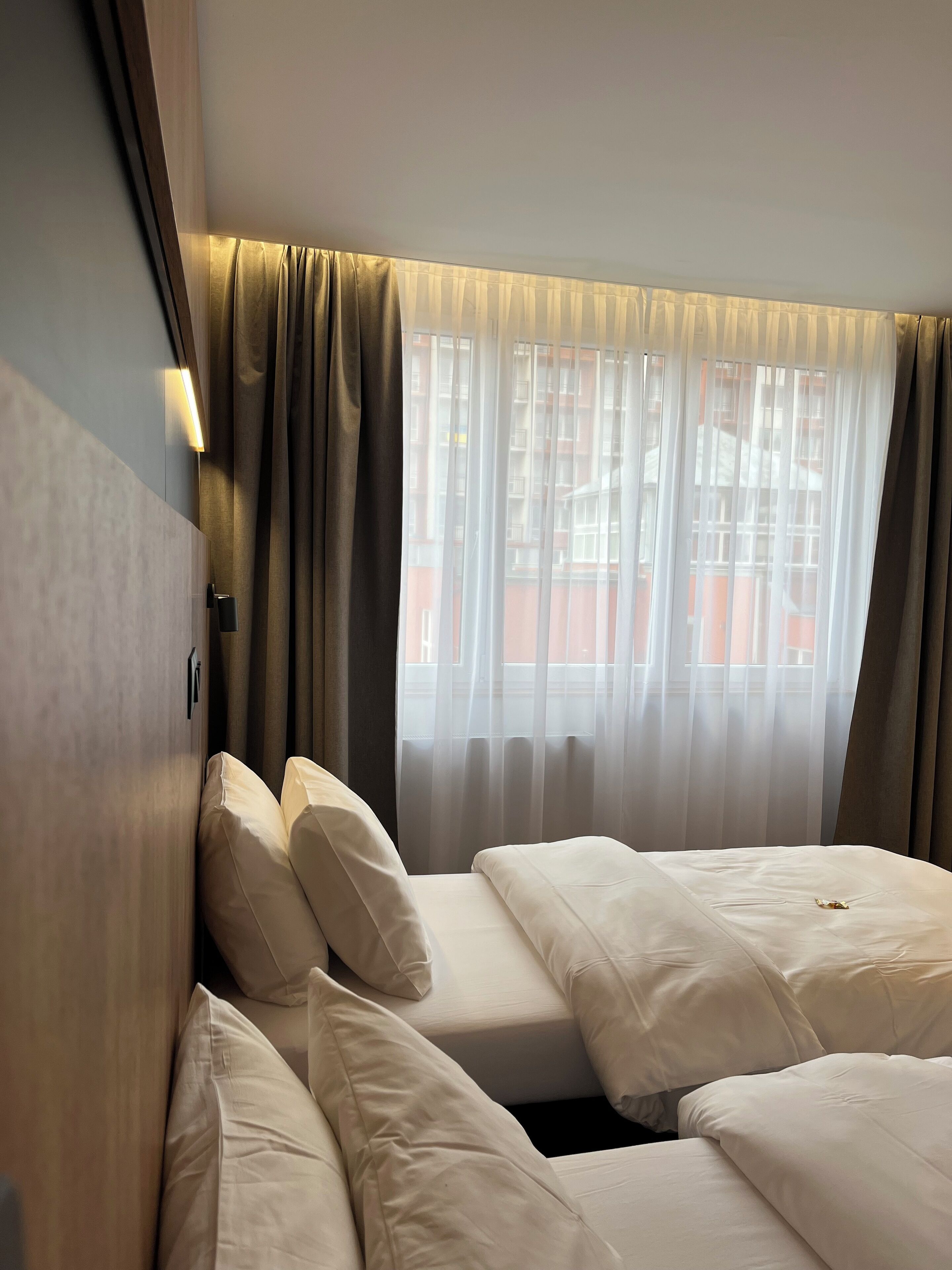 A hotel room with two beds, a window with sheer curtains, and a wall-mounted light.