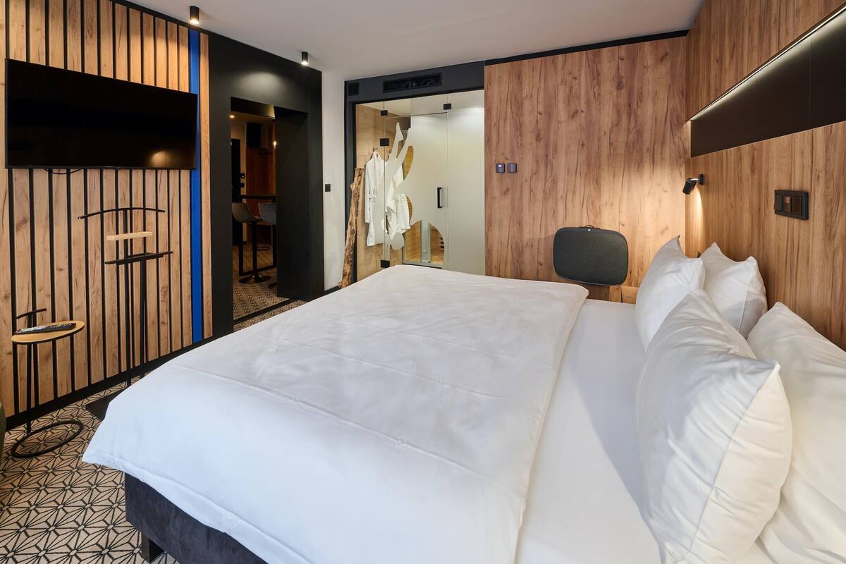 A modern hotel room with a large bed, wooden panel walls, a small seating area, and a visible bathroom.