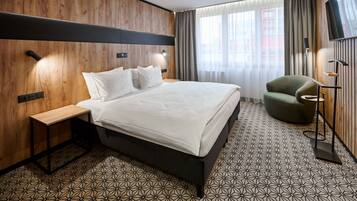 A hotel room with a bed, bedside tables, a green armchair, a TV, and a window with curtains.