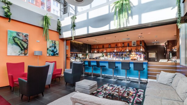 A modern bar area with a blue counter, multiple bar stools, and a variety of bottles. There are red and black chairs, a large sofa, and a coffee table in the seating area. - BEST WESTERN Hotel Vista (Ostrava)