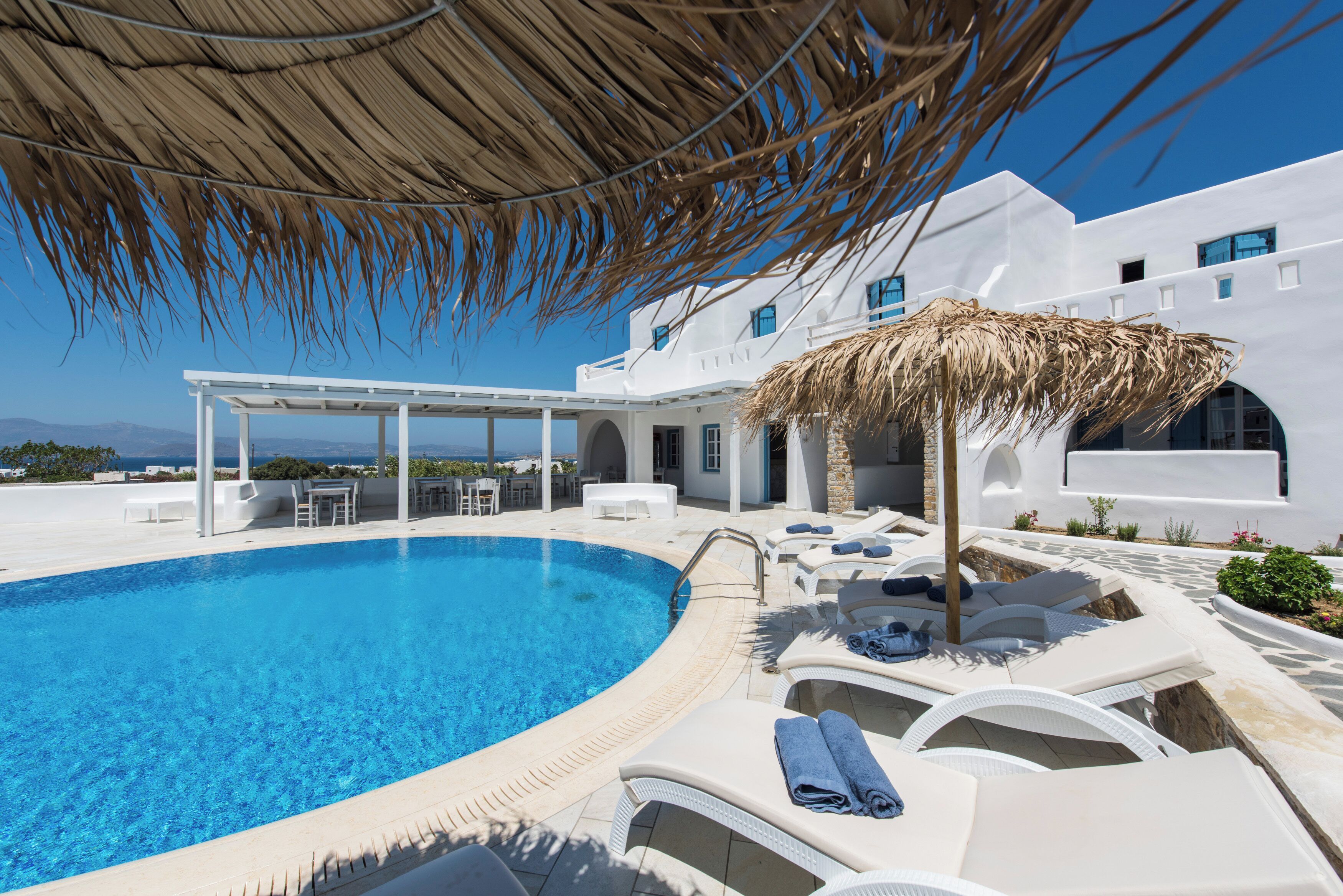 Zona de piscina con sillas reclinables y una zona de estar cubierta, rodeada de edificios blancos y un cielo azul despejado.