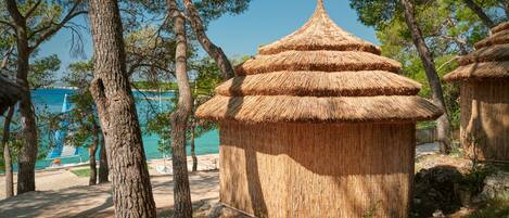 A thatched-roof hut in a natural setting with trees and a beach in the background.