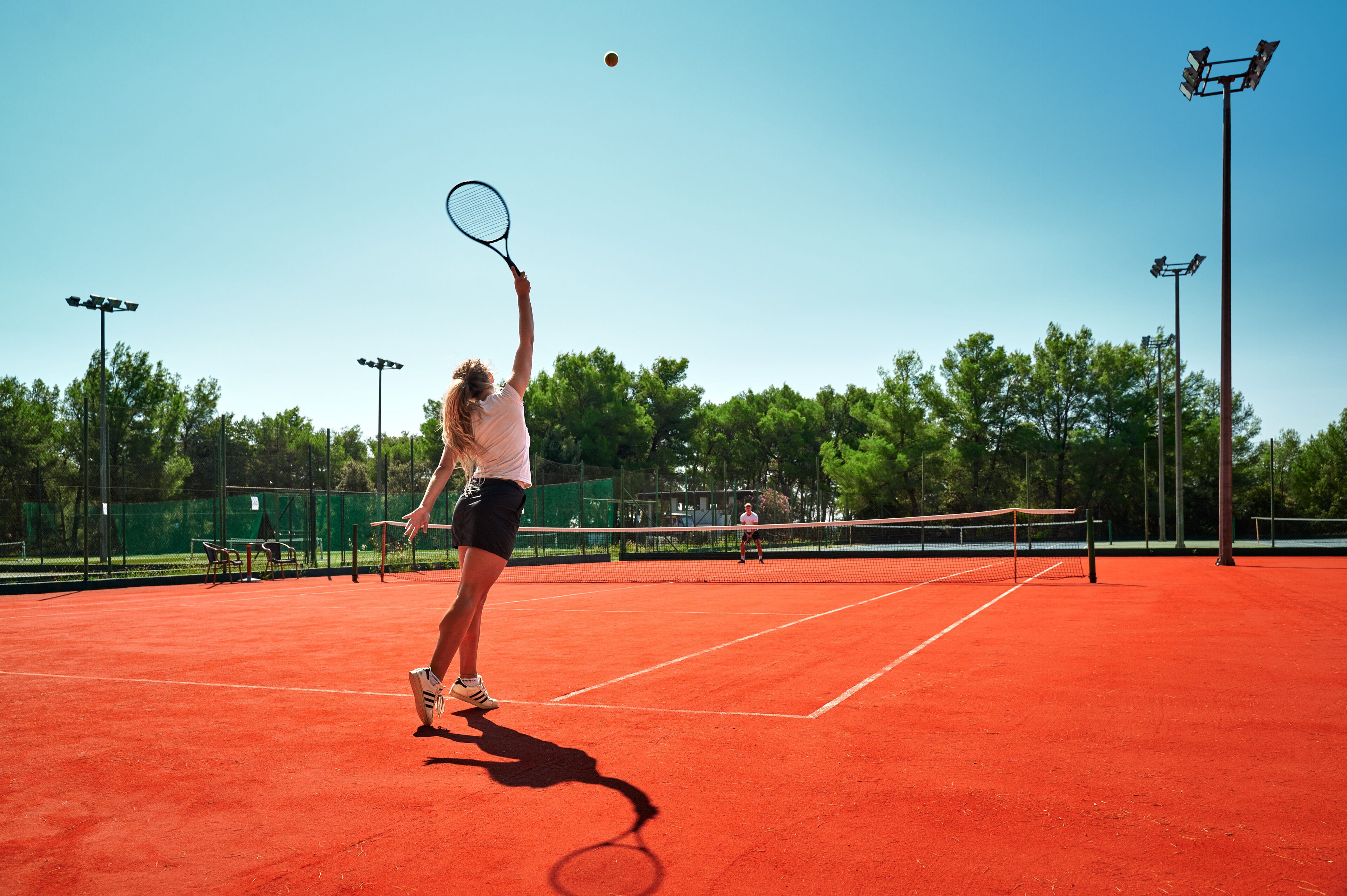 Una cancha de tenis con red, un jugador con camisa blanca y pantalon corto negro, y una raqueta de tenis.