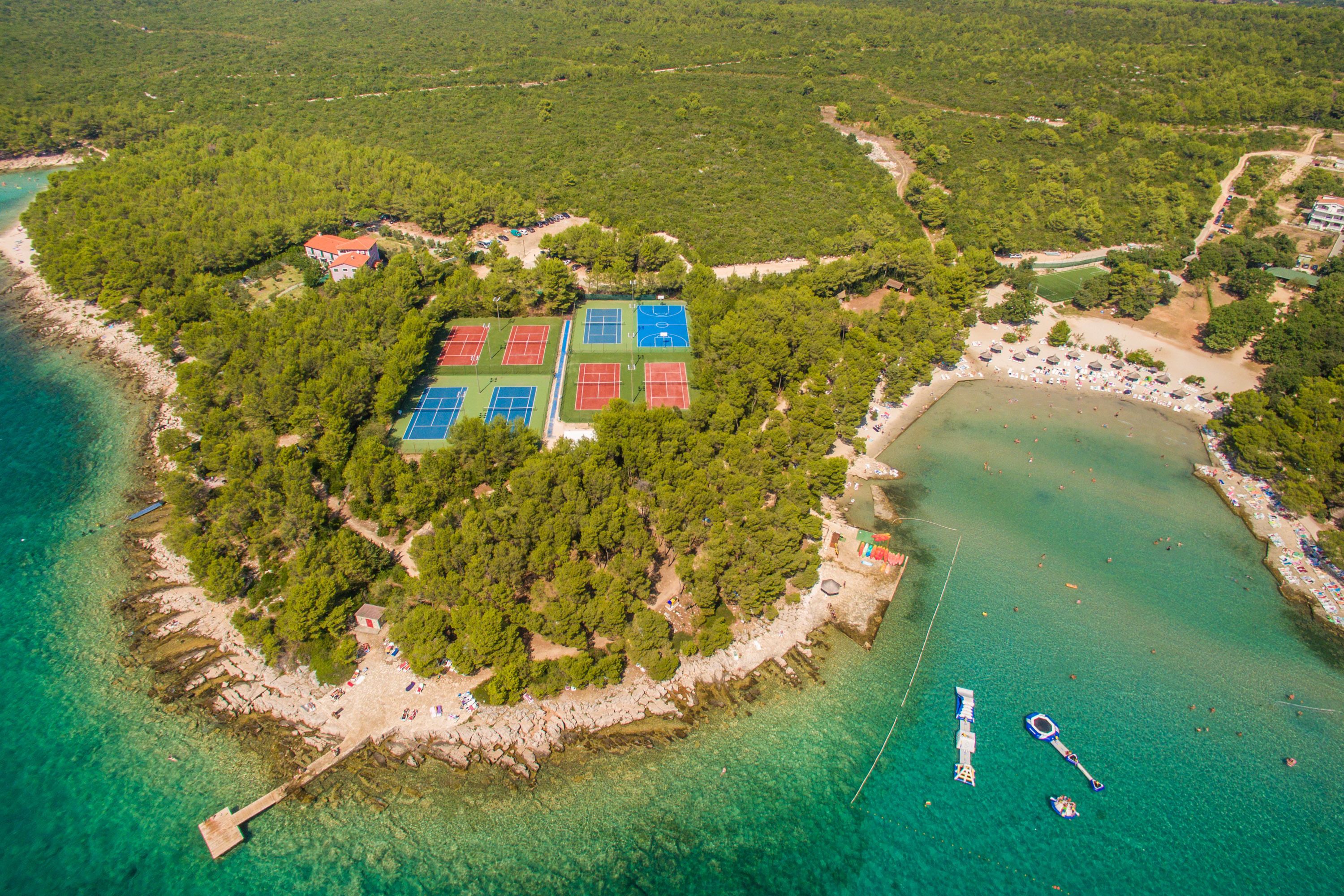 An aerial view of a coastal area with a beach, tennis courts, and a pier.