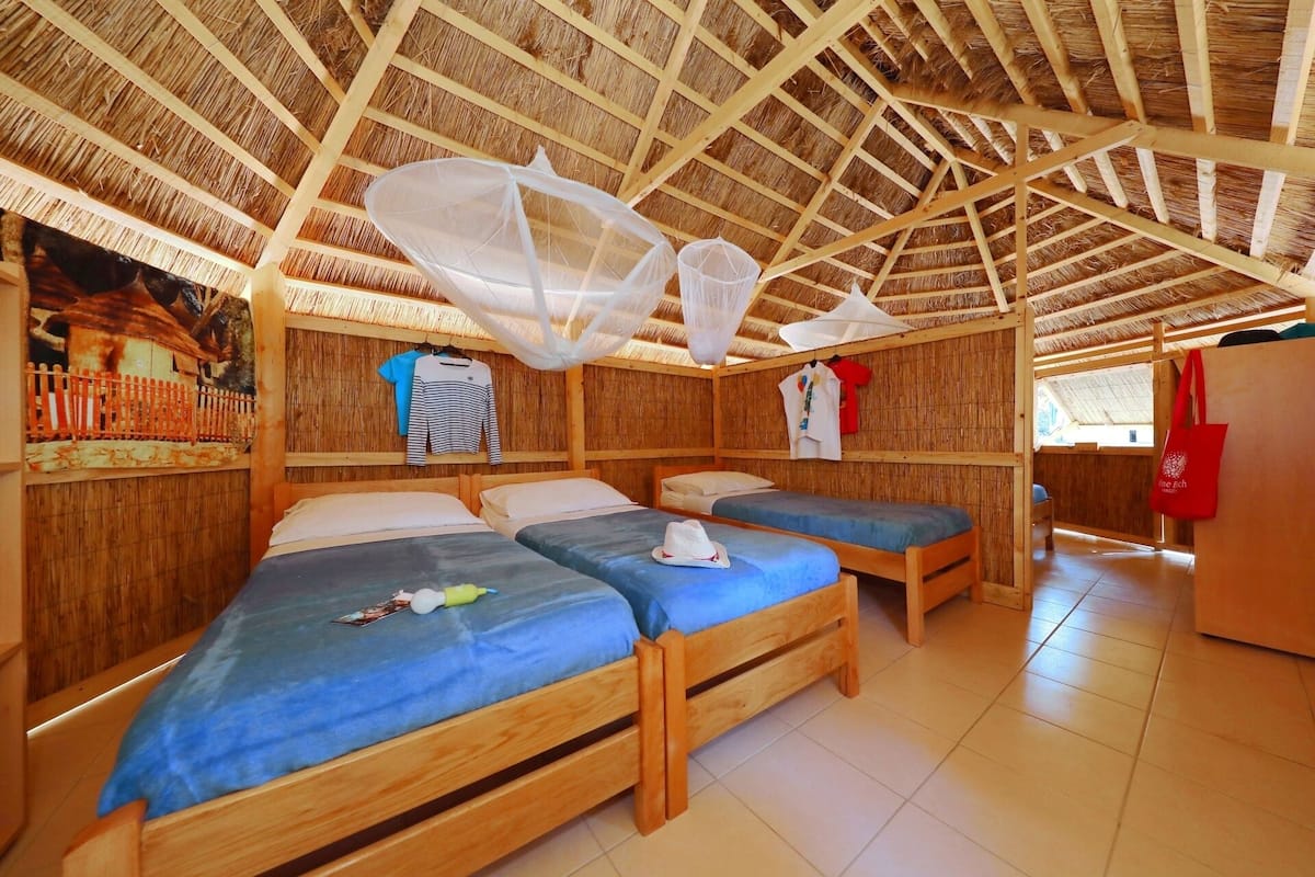 A room with two wooden beds, mosquito nets, and a red bag on a table.