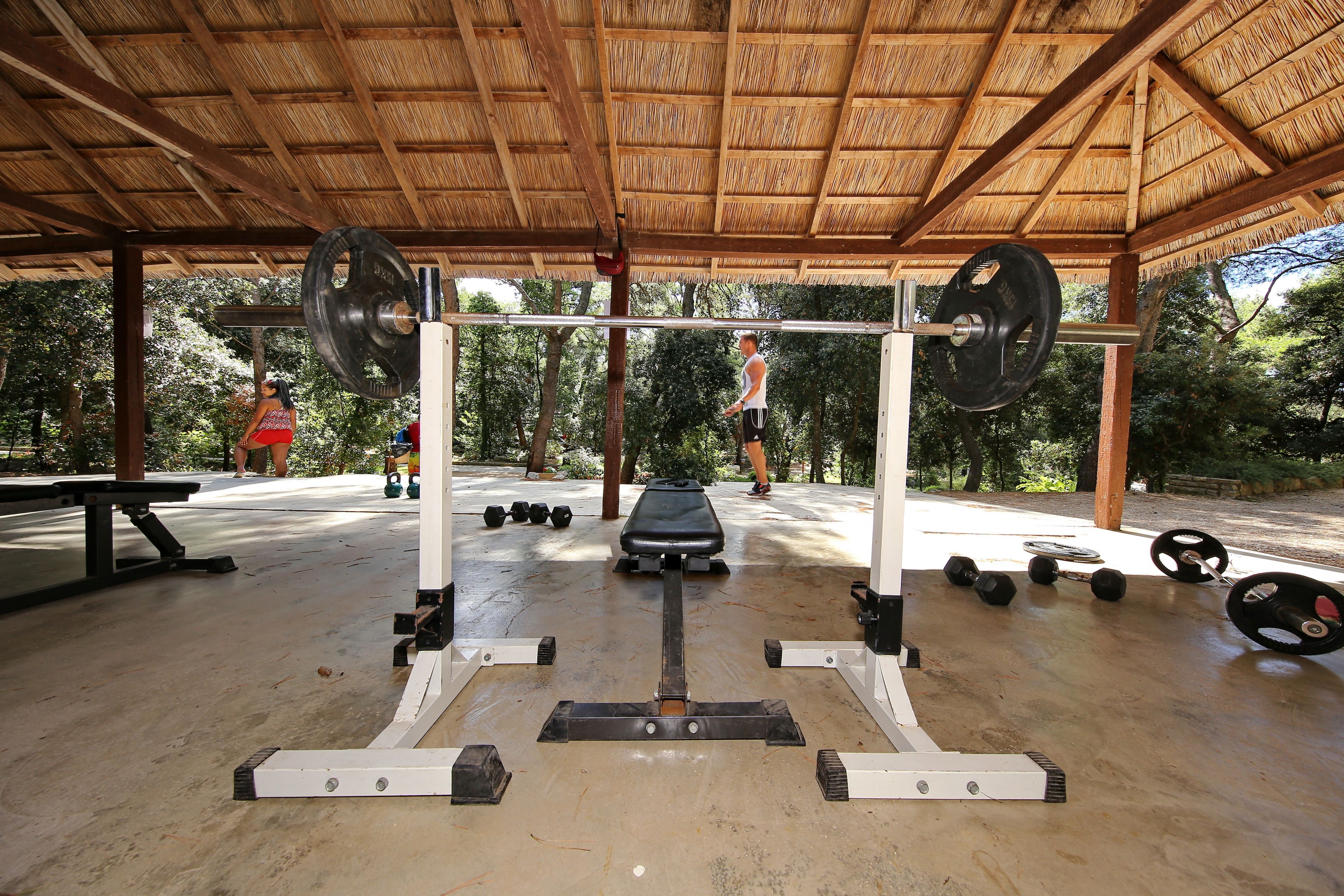Gimnasio al aire libre con techo de madera, equipo de levantamiento de pesas y colchonetas de ejercicio.