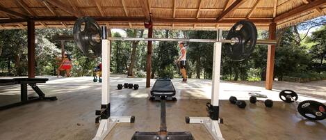 Gimnasio al aire libre con techo de madera, equipo de levantamiento de pesas y colchonetas de ejercicio.