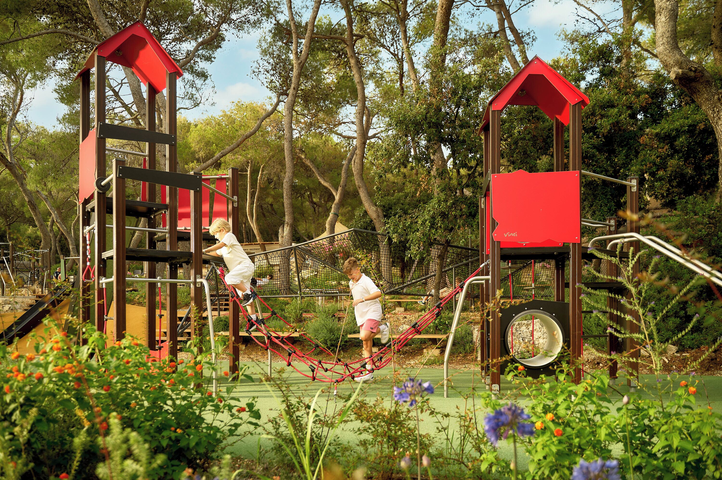 A playground with red and brown structures, surrounded by greenery and flowers.