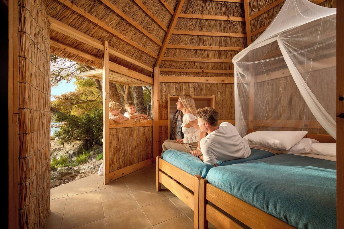 A wooden cabin with a bed, a window overlooking a rocky area, and a thatched roof.