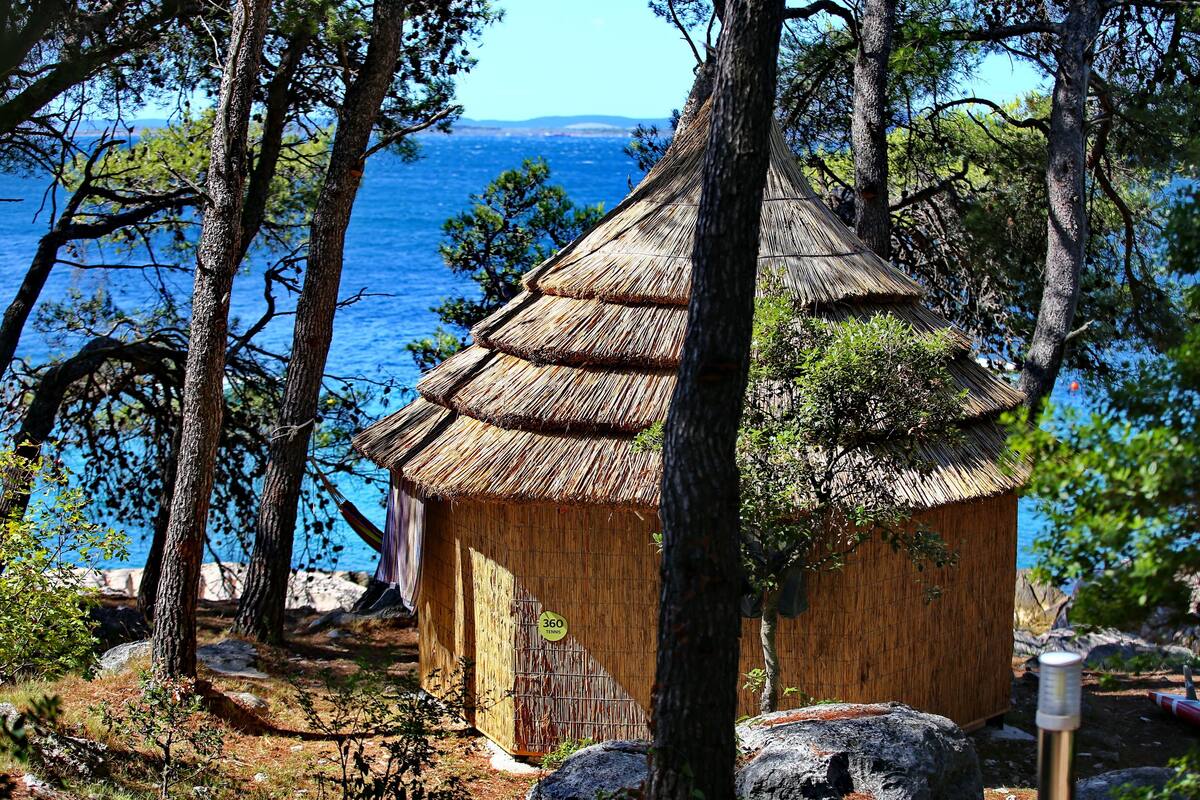 A rustic thatched-roof hut surrounded by trees, overlooking a body of water.