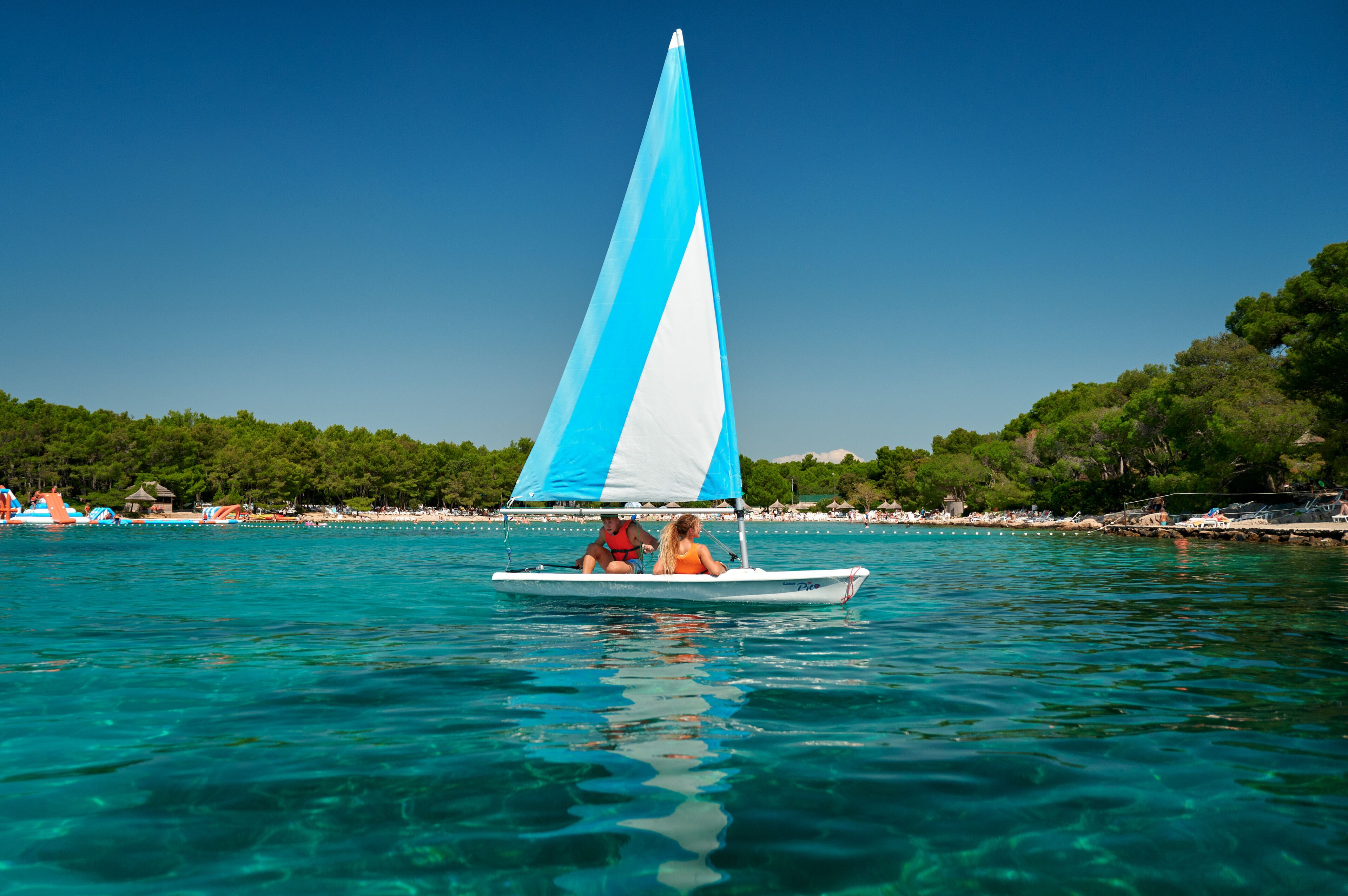 Un voilier à la voile bleue et blanche navigue sur une mer turquoise et limpide, entouré d’une plage parsemée de parasols et d’une colline boisée en arrière-plan.