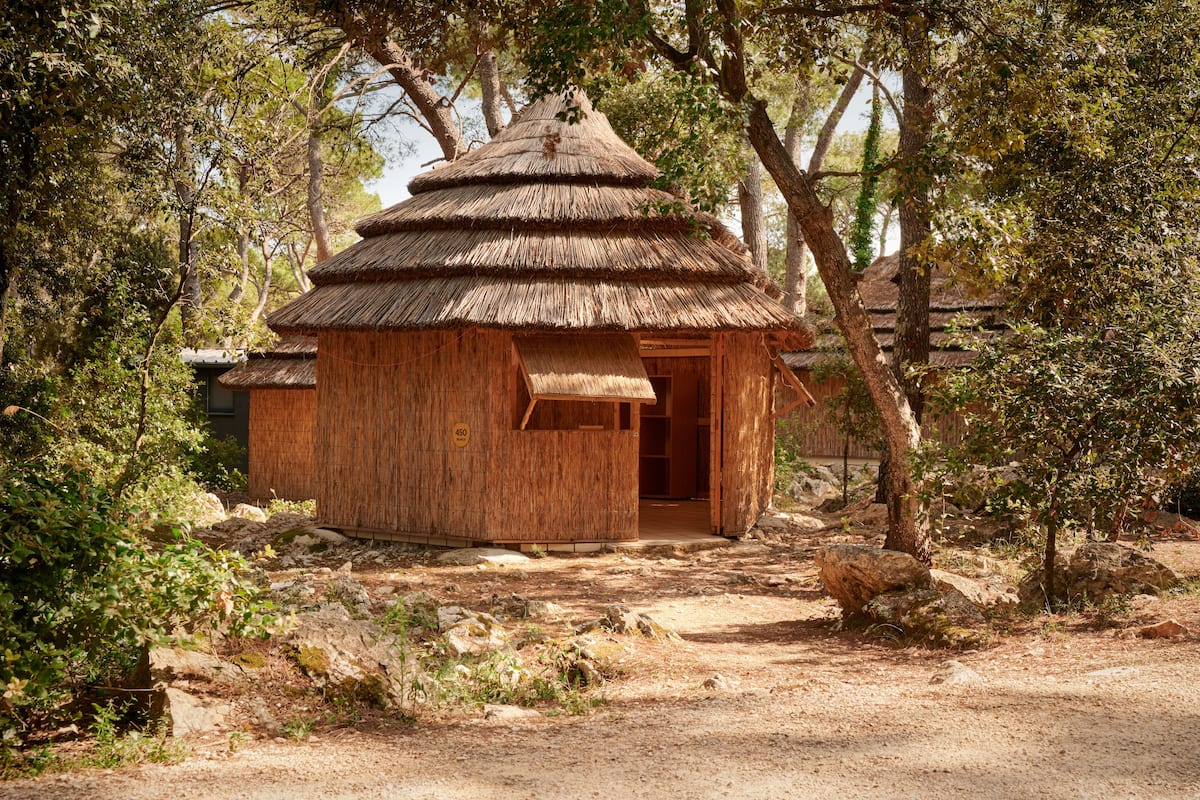 A rustic wooden hut with a thatched roof, surrounded by trees and rocks.