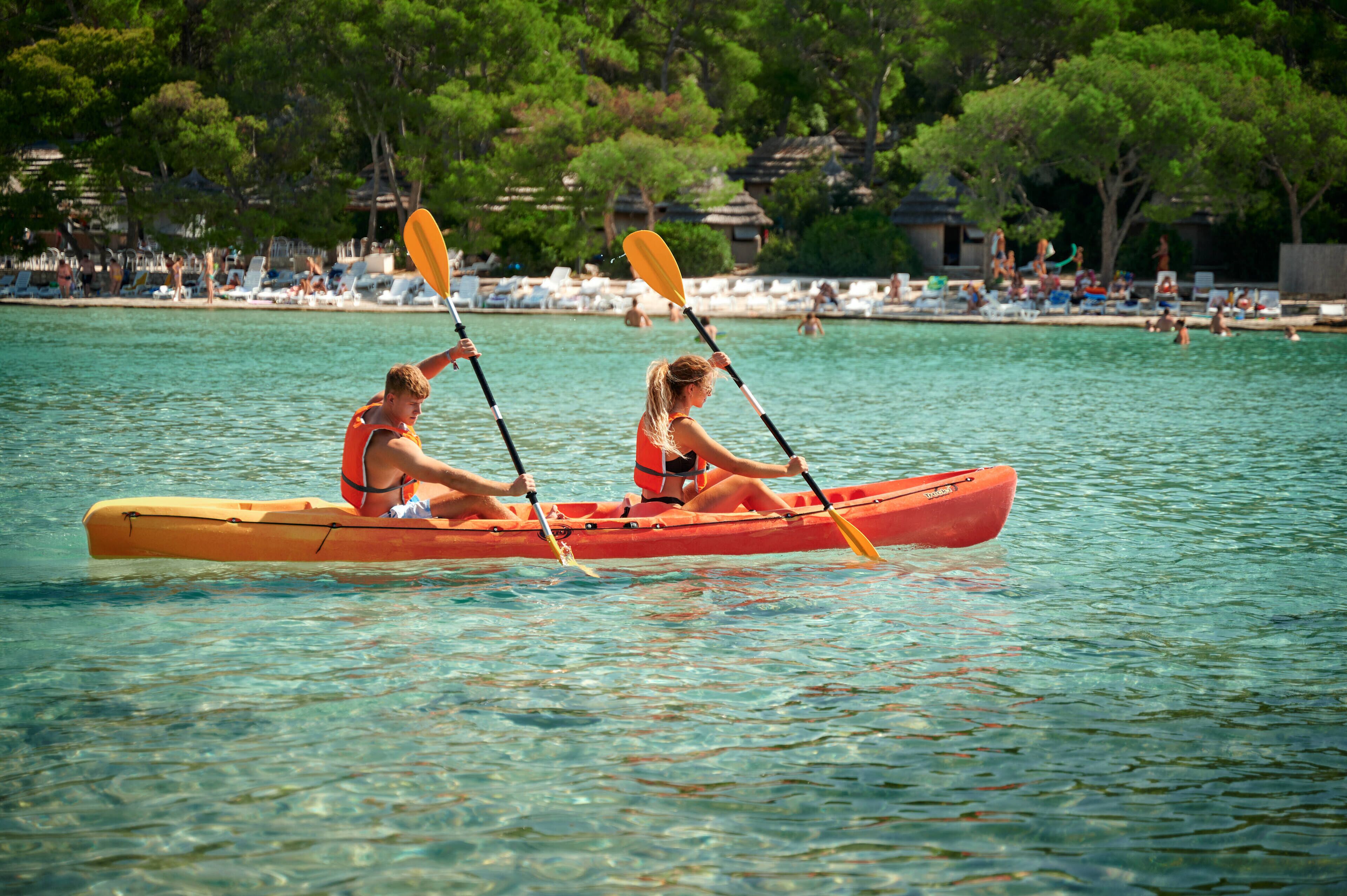 Deux personnes dans un canoë sur une étendue d’eau calme, avec une plage et des parasols en arrière-plan.