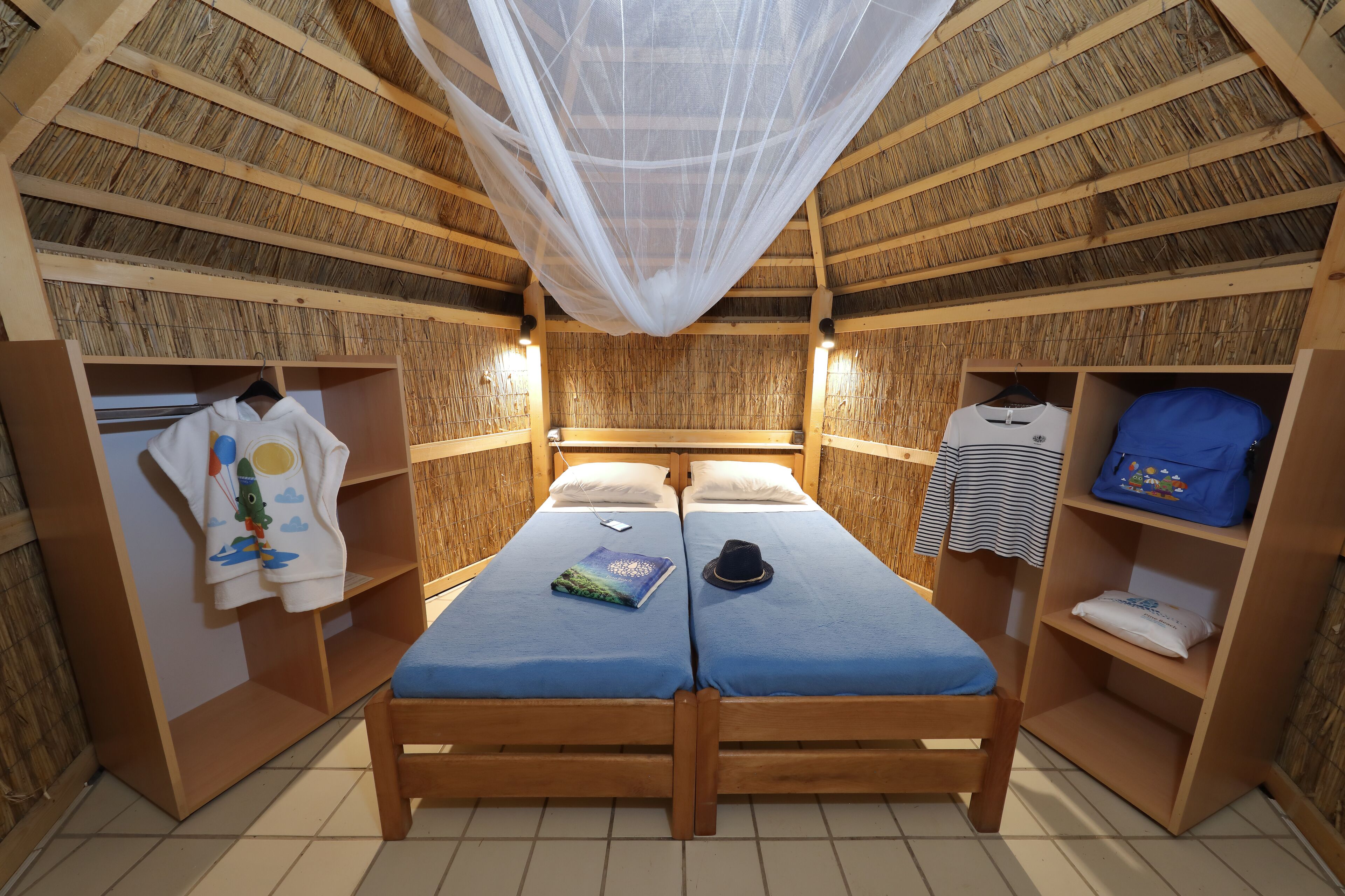 Una habitación con dos camas de madera, un mosquitero y estantes con ropa y objetos personales.