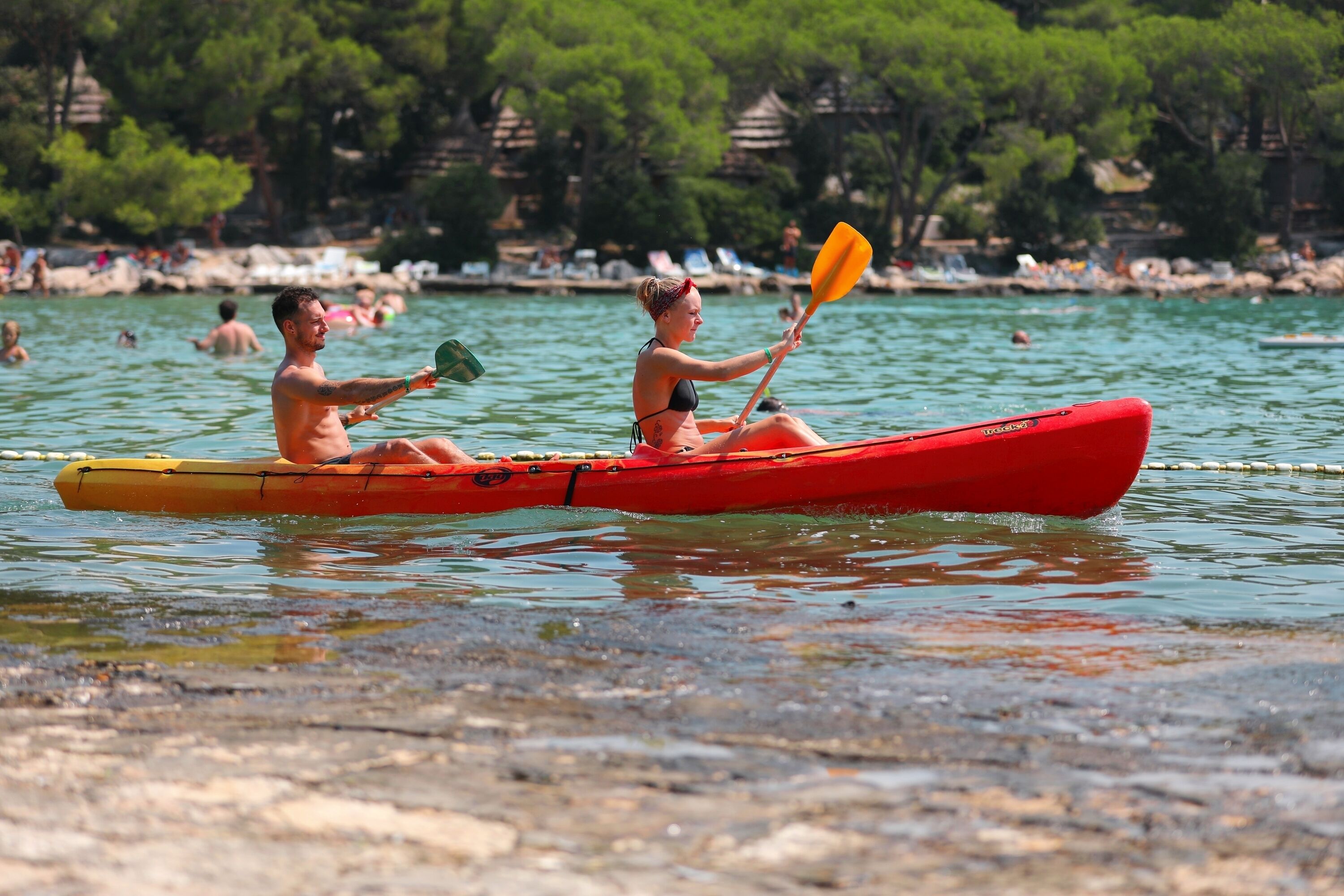 Two people in a red kayak on a calm body of water with a beach and trees in the background.