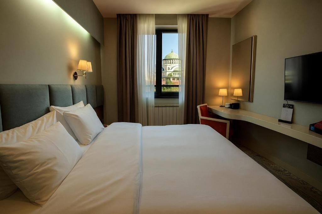 A hotel room with a large bed, a desk with a chair, a television, and a view of a building through a window.