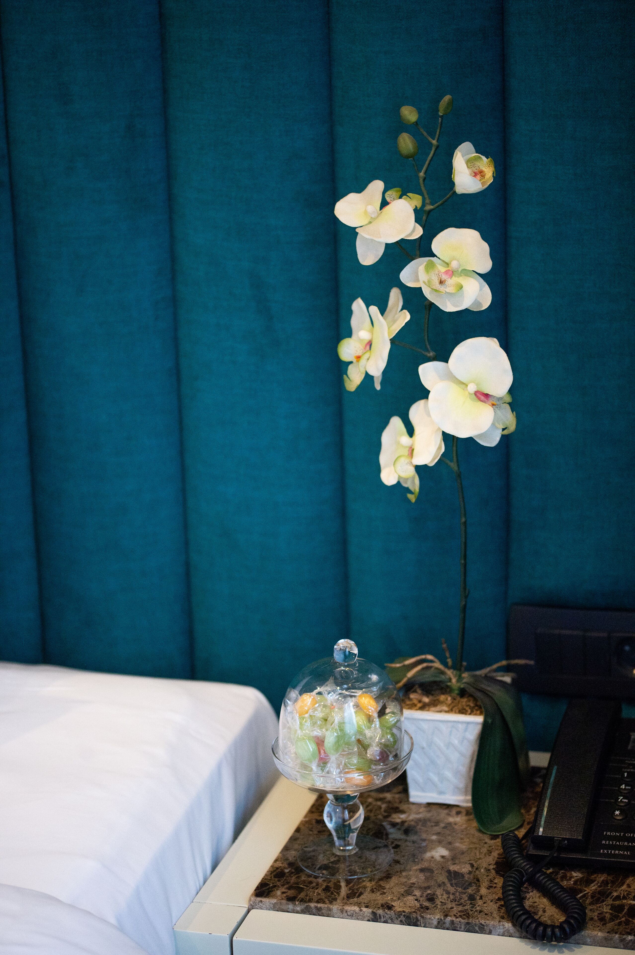 Une chambre d’hôtel avec un lit, un saladier en verre rempli de fruits, une plante en pot et un téléphone posé sur une surface en marbre.
