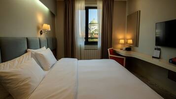 Habitación de hotel con una cama grande, un escritorio con silla, un televisor y una ventana con vista a un edificio.